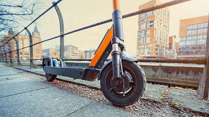 MASTERLOCK 66268700 Street Cuff Ein grauer Roller mit orangen Akzenten steht auf einem Bürgersteig. Im Hintergrund sind Gebäude und eine Metallbarriere zu sehen.