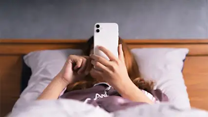 Eine Person liegt im Bett und hält ein weißes Smartphone hoch. Das Bett hat ein hölzernes Kopfende und weiße Kissen.