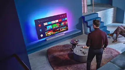 Ein Wohnzimmer mit einem großen Fernseher an der Wand, einem Couchtisch und zwei Personen im Hintergrund.