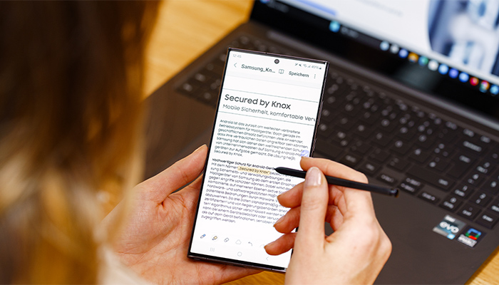 Eine Frau schreibt mit einem Stift auf Ihrem Smartphone-Display, im Hintergrund steht ein aufgeklappter Laptop.