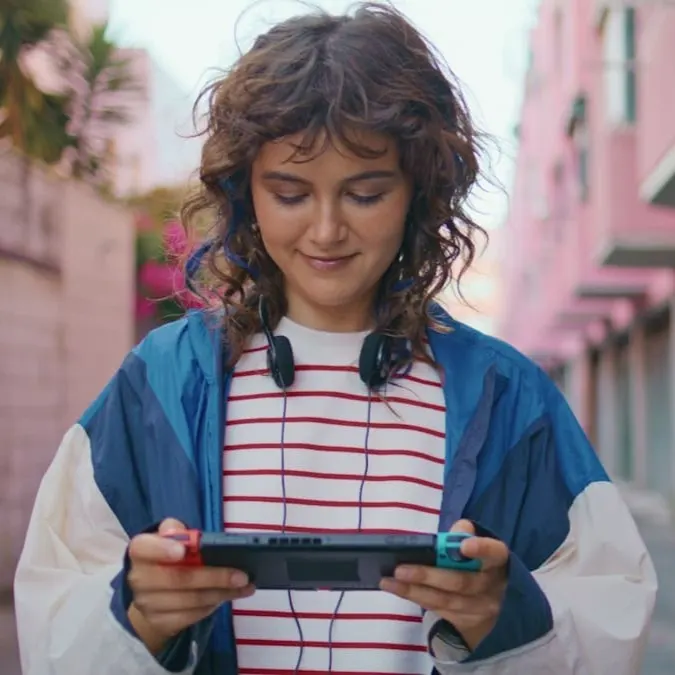 Junge Frau steht draußen in einer Gasse und spielt entspannt auf einer Nintendo Switch mit roten und blauen Controllern.