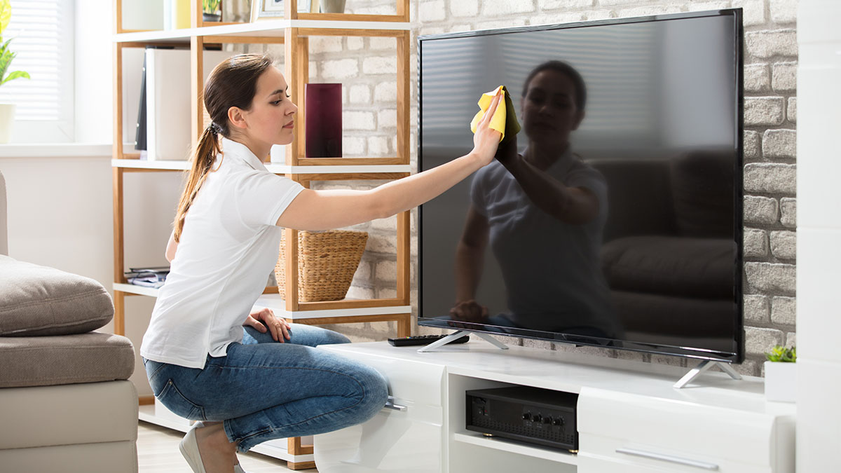 Frau putzt mit einem gelben Tuch den Bildschirm eines großen Fernsehers im Wohnzimmer.