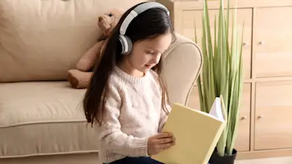 Ein Mädchen mit Kopfhörern liest ein gelbes Buch. Im Hintergrund sind ein Sofa, ein Teddybär und eine Pflanze.