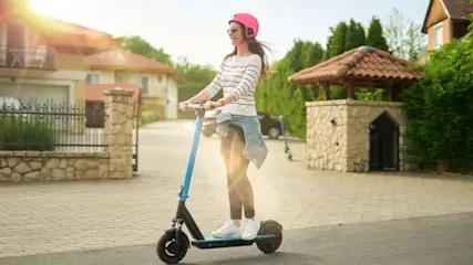 Eine Frau mit rosa Helm fährt einen blauen Roller auf einer gepflasterten Straße. Im Hintergrund sind Häuser und Grün zu sehen.