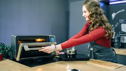 Eine Frau öffnet einen Ofen in einer Küche. Auf der Arbeitsfläche stehen ein Kochfeld, ein Topf und ein Schneidebrett.