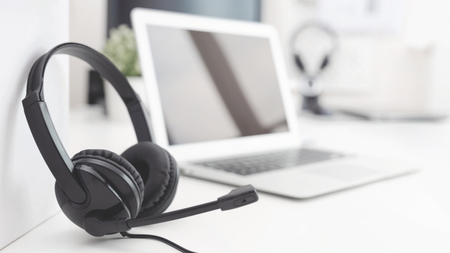 Schwarzes Headset mit Mikrofon vor einem weißen Laptop auf einem weißen Tisch. Ein weiteres Headset ist im Hintergrund zu sehen. (Adobe Stock, Datei Nr.:265958720)