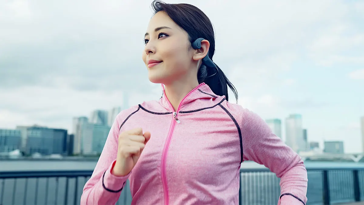 Frau in pinker Sportjacke joggt mit Open-Ear-Kopfhörern am Wasser, im Hintergrund die Skyline einer Stadt.