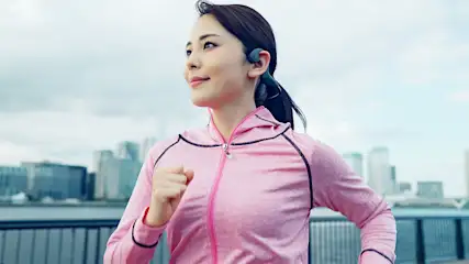 Frau in pinker Sportjacke joggt mit Open-Ear-Kopfhörern am Wasser, im Hintergrund die Skyline einer Stadt.