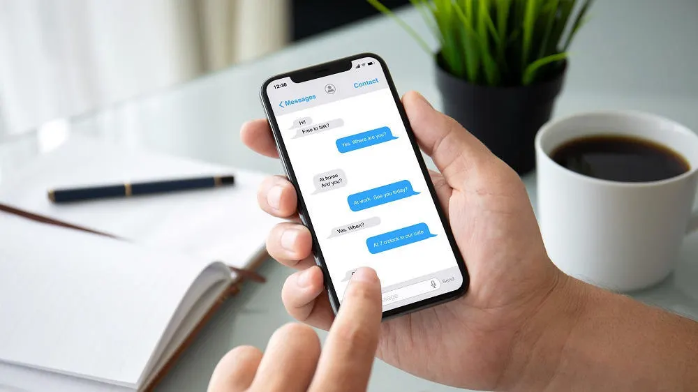 Eine Hand hält ein Smartphone mit geöffnetem Chat. Auf einem Tisch liegen ein Notizbuch, ein Stift, eine Kaffeetasse und eine Pflanze.