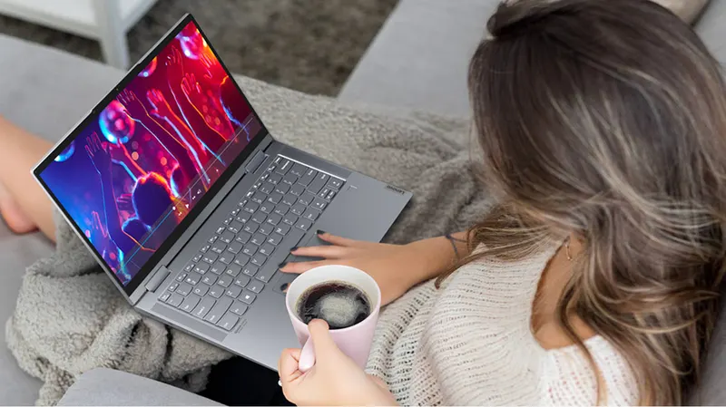 Eine Person sitzt mit einem Laptop und einer Tasse Kaffee auf einem grauen Sofa unter einer grauen Decke.