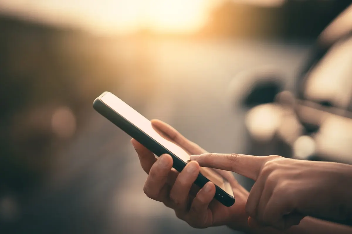 Eine Hand hält ein Smartphone mit schwarzem Rahmen und tippt darauf. Im Hintergrund ist ein Auto zu sehen.