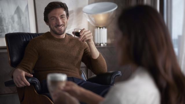 Ein Mann im braunen Pullover sitzt in einem Sessel und hält ein Weinglas. Eine Frau im weißen Oberteil ist im Vordergrund.