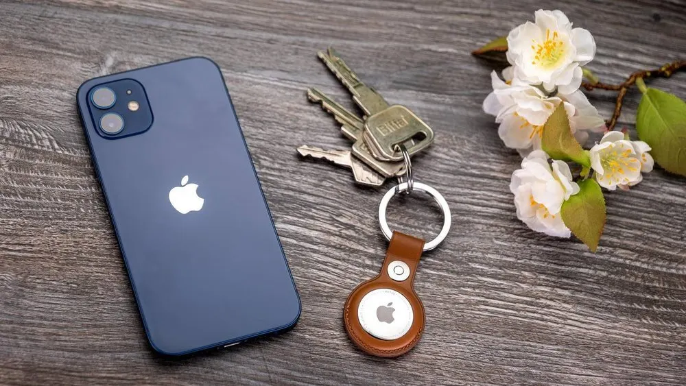 Blaues Smartphone, Schlüsselbund mit AirTag und weiße Blüten auf einem Tisch mit Holzstruktur.
