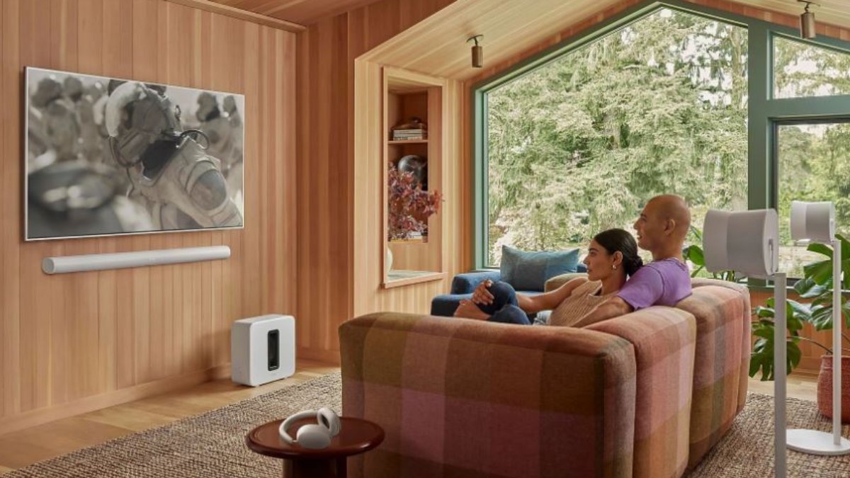 Wohnzimmer mit Holzvertäfelung, Paar auf Sofa vor TV, Soundbar, Subwoofer und Lautsprechern. Fenster mit Blick auf Bäume.