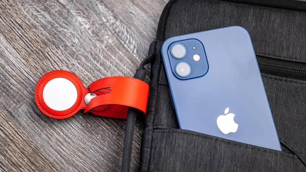 Ein blaues Smartphone steckt in einer schwarzen Tasche. Ein roter AirTag-Halter liegt auf einer grauen Holzoberfläche.