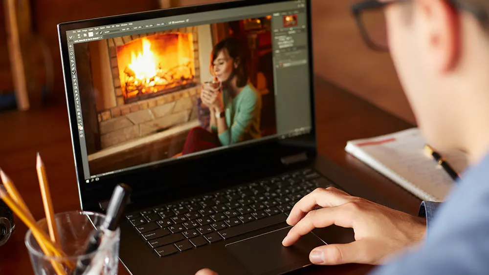 Person arbeitet am Laptop. Auf dem Bildschirm ist eine Frau vor einem Kamin zu sehen. Notizbuch und Stifte liegen auf dem Tisch.
