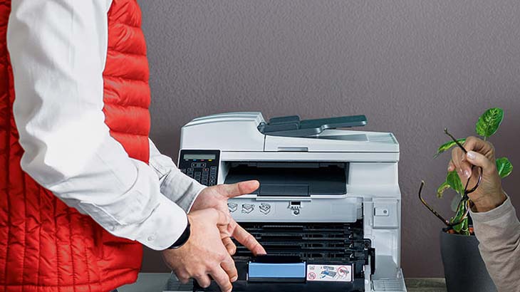 Eine Person wechselt eine Tonerkartusche in einem weißen Drucker, während eine andere Person eine Brille über einer Pflanze hält.