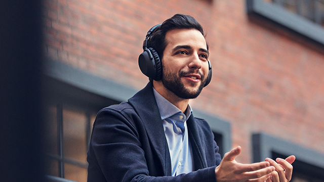 Ein Mann mit Kopfhörern, Anzug und Hemd gestikuliert vor einem Backsteingebäude.