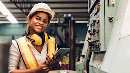 Eine Frau mit Schutzhelm und gelben Kopfhörern hält ein Tablet in einer Fabrik neben einer Maschine mit Knöpfen.