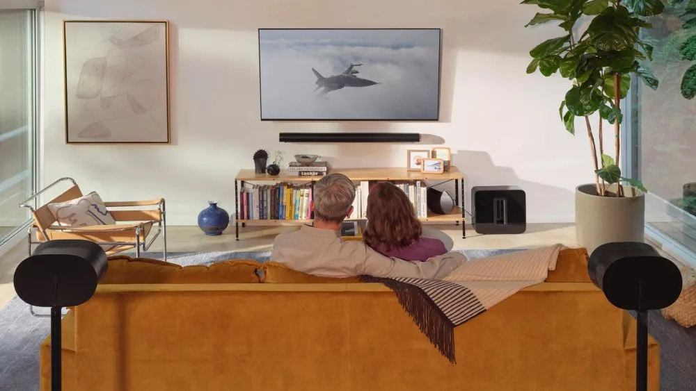 Paar sitzt auf einem gelben Sofa, vor ihnen ein Fernseher mit einem Flugzeug. Regale, Stuhl und Pflanze im Hintergrund.