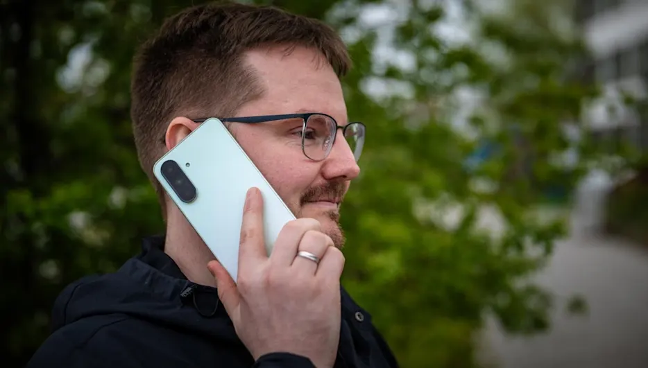 Mann mit Brille und Bart telefoniert mit hellblauem Smartphone vor grünem Hintergrund. Er trägt eine schwarze Jacke.