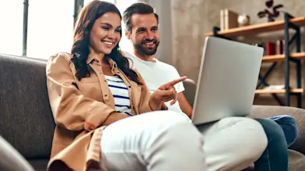 Ein Paar sitzt auf einem grauen Sofa und schaut auf einen Laptop. Der Mann zeigt auf den Bildschirm. Im Hintergrund ist ein Regal.