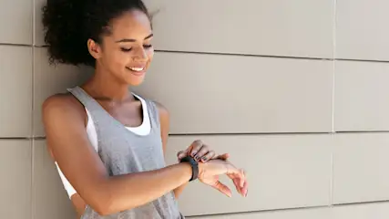 Eine Frau im Sporttrikot schaut auf eine schwarze Smartwatch am Handgelenk vor einer hellen Wand.
