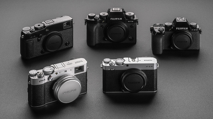Fünf Fujifilm-Kameras unterschiedlicher Modelle liegen auf einer grauen Oberfläche. Die Kameras sind schwarz, silber und haben Objektivdeckel.