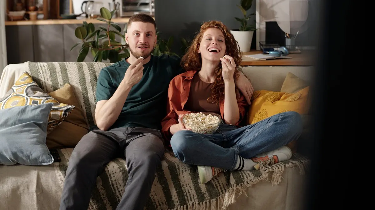 Ein Paar sitzt auf einem Sofa mit Streifen und isst Popcorn vor einem dunklen Bildschirm. Kissen und Pflanzen sind im Hintergrund.