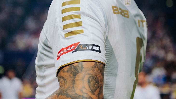 Ein Mann trägt ein weißes Trikot mit goldenen Streifen und den Logos von Media Markt und Saturn auf dem Ärmel. Eine Tätowierung ist sichtbar.