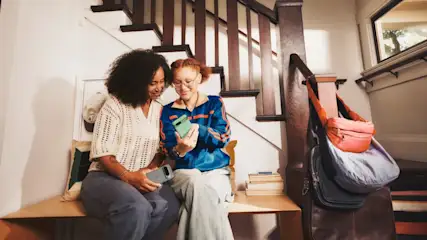 Zwei Personen sitzen auf einer Bank und betrachten ein Smartphone vor einer Treppe. Eine Tasche hängt an der Treppe.