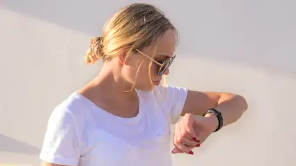 Frau mit Sonnenbrille und weißem T-Shirt blickt auf ihre schwarze Armbanduhr vor einer hellen Wand.