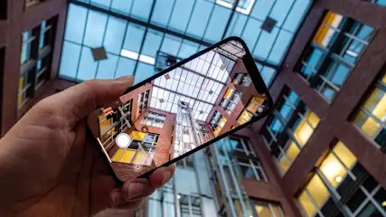 Eine Hand hält ein Smartphone und fotografiert ein Gebäude mit Glasdach und Aufzugsschacht.