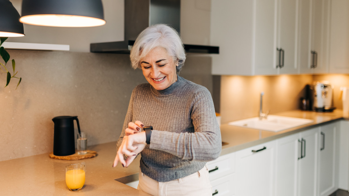 Eine ältere Frau steht in einer Küche und blickt lächelnd auf die Smartwatch an ihrem Handgelenk.