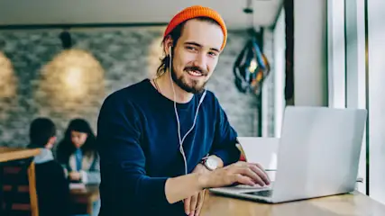 Ein Mann mit orangefarbener Mütze tippt auf einem Laptop, Kopfhörer im Ohr. Hintergrund: Andere Personen sitzen an Tischen.