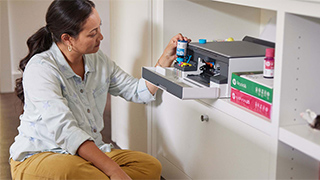 Frau sitzt vor Drucker auf weißem Regal mit Farbdruckflaschen. Grüne und rote Boxen sind daneben gestapelt.