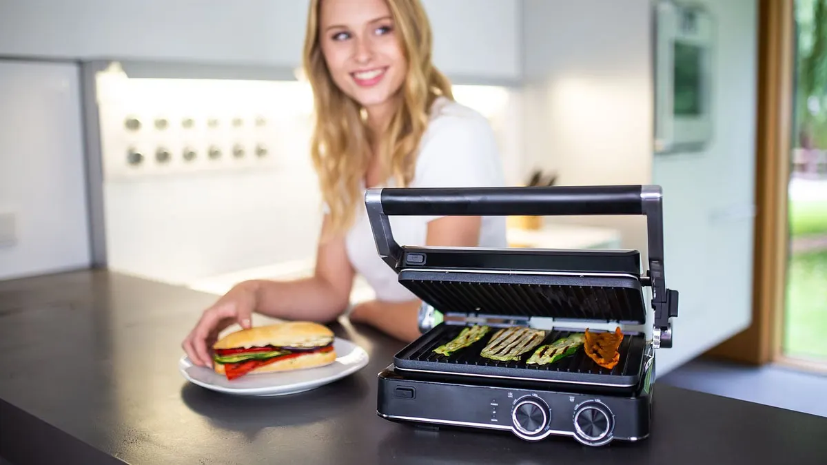 Eine Frau hat einen Teller mit einem Baguette mit Grillgemüse vor sich, daneben steht der geöffnete Kontaktgrill KOENIC KCG 2020M.