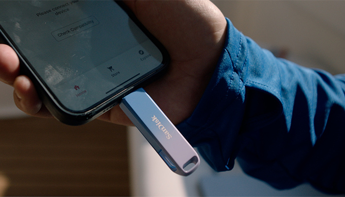 Hand hält iPhone, blauer SanDisk‑USB‑Stick eingesteckt; App‑Bildschirm sichtbar.