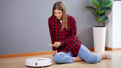 Eine Frau sitzt auf einem Holzboden mit einem Handy, neben einem Staubsaugerroboter und einer Zimmerpflanze.