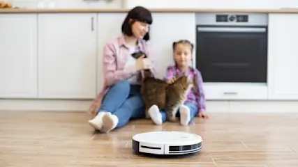 Auf dem Boden sitzt eine Frau mit Kind und Katze. Ein Staubsaugerroboter steht im Vordergrund. Im Hintergrund sind Küchenschränke.