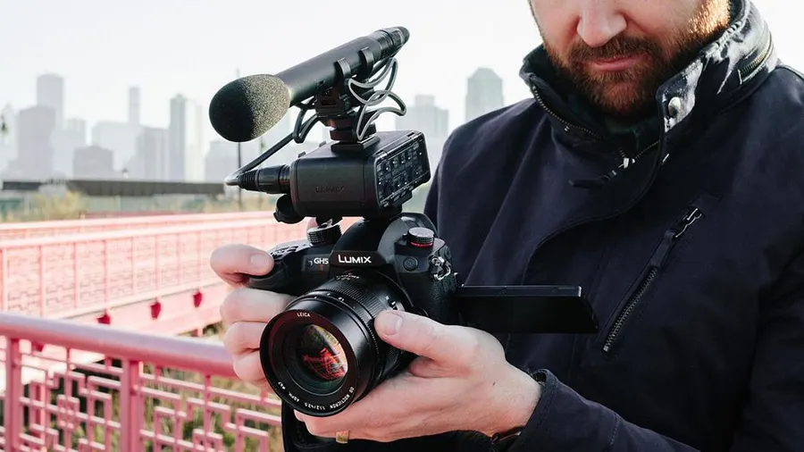 Ein Mann hält eine schwarze Lumix-Kamera mit Mikrofon vor einer Stadtkulisse und einem roten Geländer.