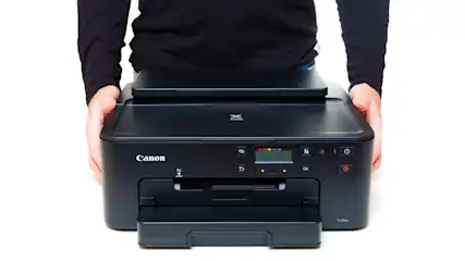 Eine Person in schwarzer Kleidung hält einen schwarzen Canon-Drucker mit Bedienfeld und Papiereinzug vor einem weißen Hintergrund.
