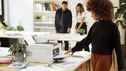 Eine Frau nimmt Papier aus einem weißen Drucker in einem Büro. Zwei Personen stehen an einem Tisch im Hintergrund.