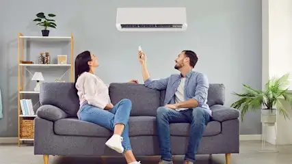Eine Frau und ein Mann sitzen auf einem Sofa. Der Mann bedient mit einer Fernbedienung eine Klimaanlage.