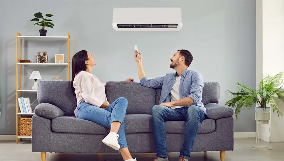 Eine Frau und ein Mann sitzen auf einem Sofa. Der Mann bedient mit einer Fernbedienung eine Klimaanlage.