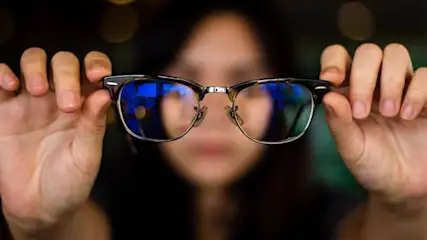 Eine Person hält eine Brille mit silbernem Rahmen und blauen Gläsern vor ihr Gesicht. Der Hintergrund ist dunkel.