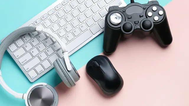 Eine weiße Tastatur, graue Kopfhörer, eine schwarze Maus und ein schwarzer Gamecontroller liegen auf einem zweifarbigen Hintergrund.