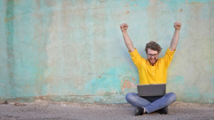 Mann im gelben Hemd sitzt im Schneidersitz auf dem Boden draußen. Ein Laptop ist auf seinem Schoss und er reißt die Arme hoch als Zeichen der Freude.