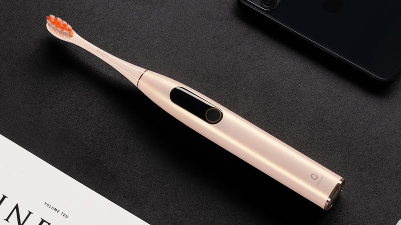 Eine goldene elektrische Zahnbürste liegt auf einer schwarzen Oberfläche, neben einem Telefon und einem weißen Dokument.