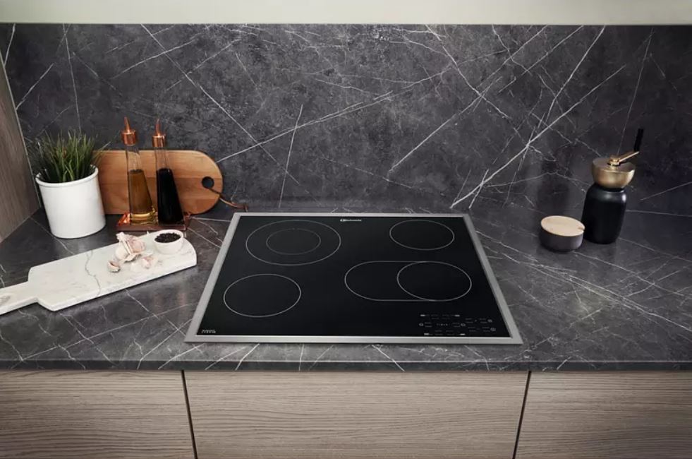 Küchenszene mit schwarzem Kochfeld, Kräutern im Topf, Schneidebrett und Marmor-Arbeitsplatte vor gemustertem Hintergrund.